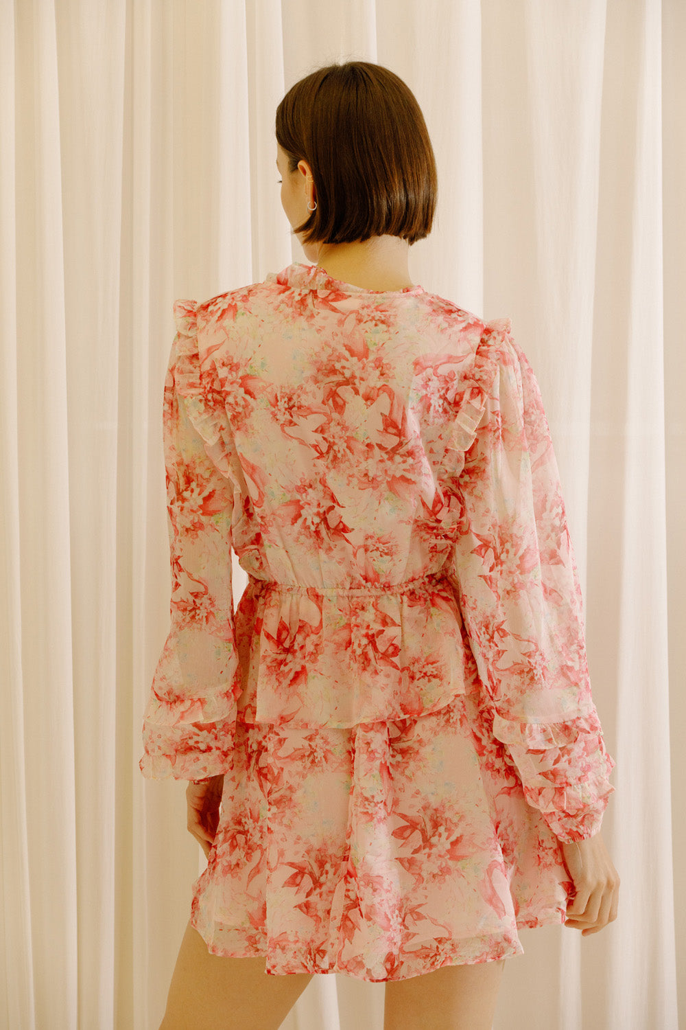 Back view of a woman wearing a pink and cream dress with floral print against a white curtain background