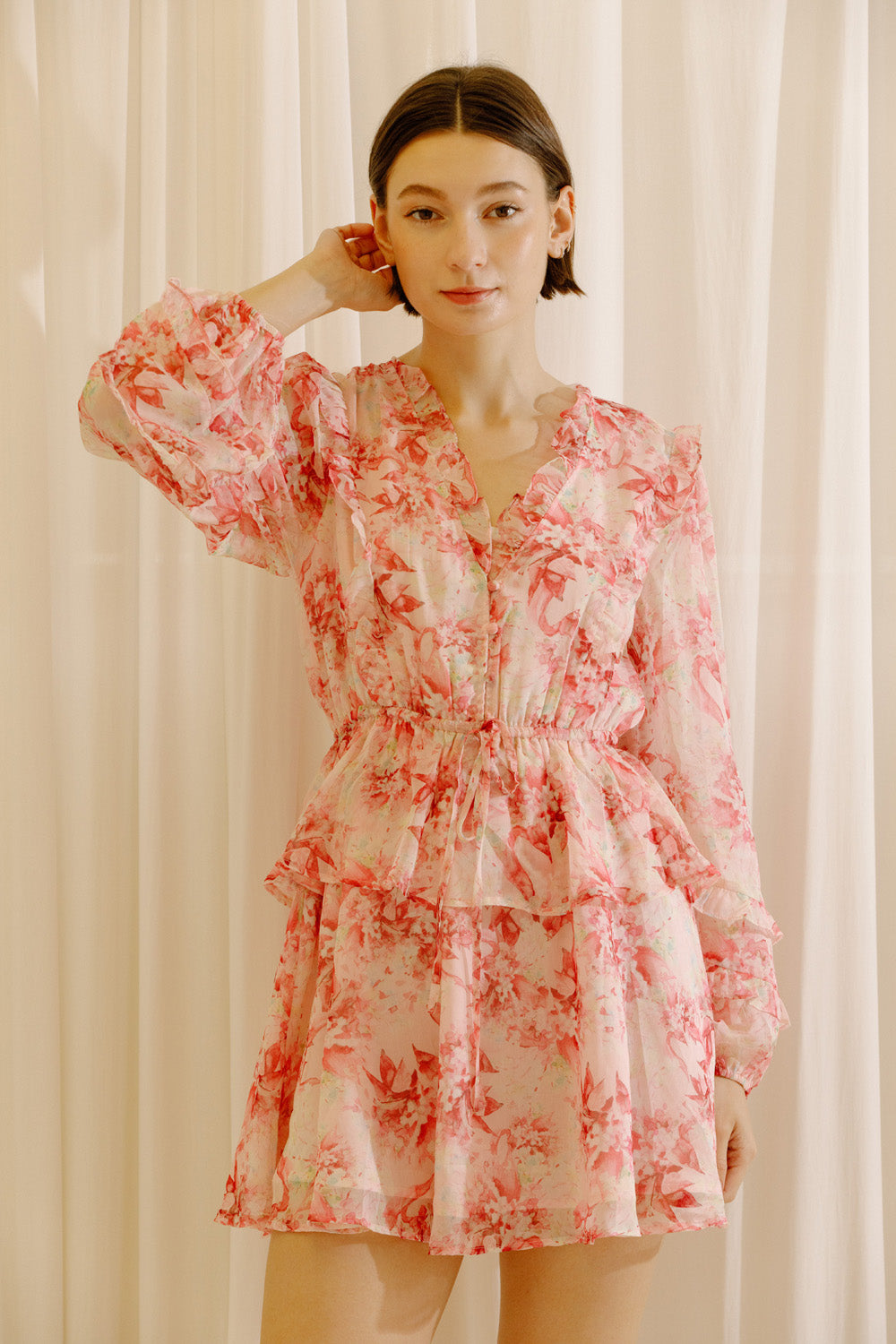 Woman wearing a pink and cream dress with floral print against a white curtain background