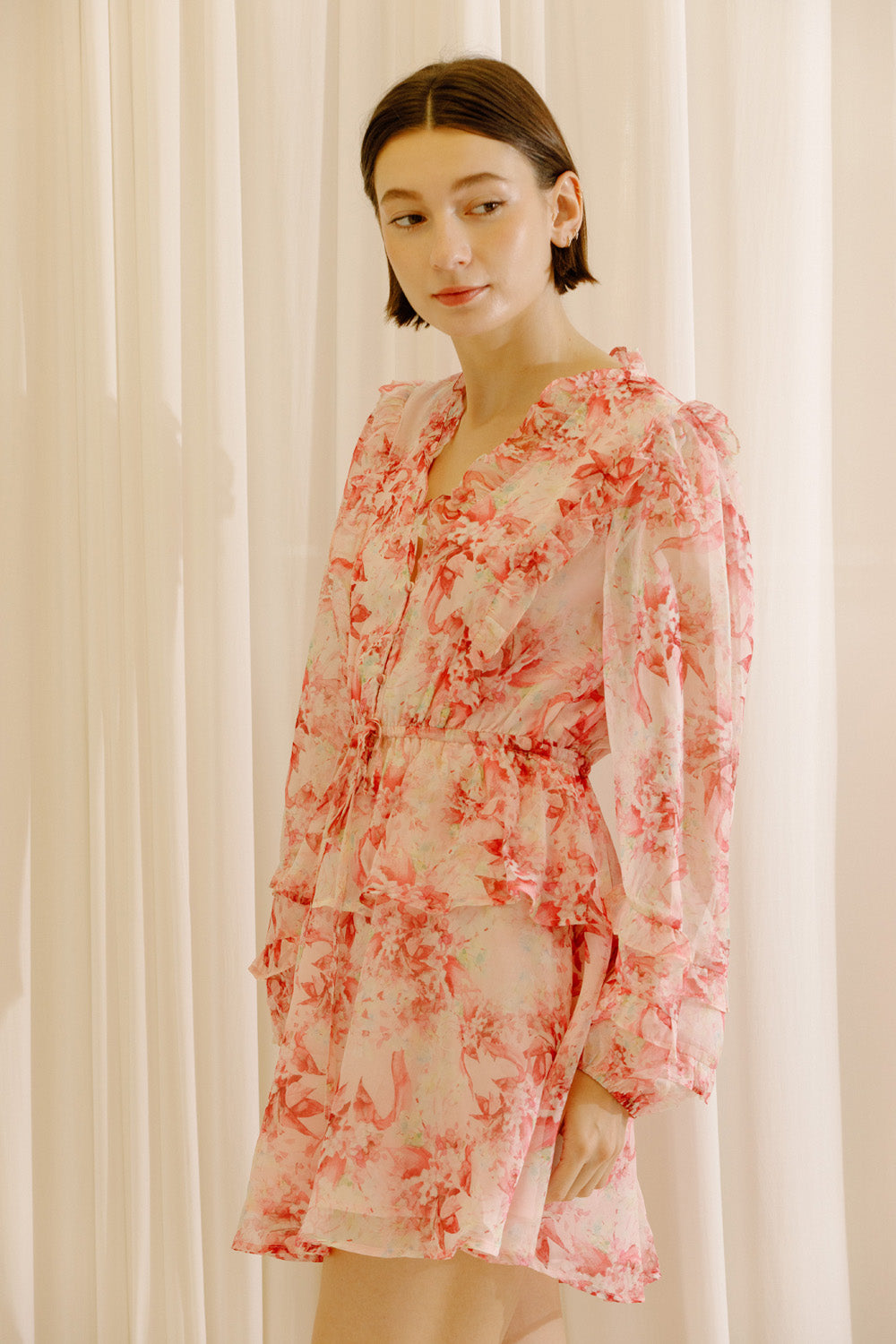 Side view of a woman wearing a pink and cream dress with floral print against a white curtain background