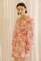Side view of a woman wearing a pink and cream dress with floral print against a white curtain background