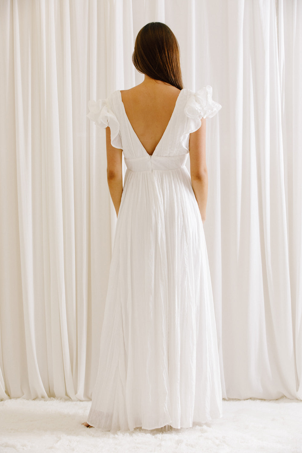 Back view of a woman wearing a white maxi dress standing against a white curtain
