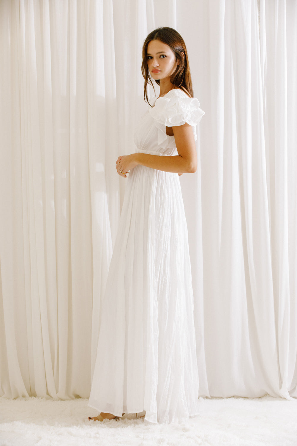 Side view of a woman wearing a white maxi dress standing against a white curtain