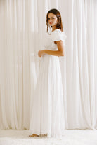 Side view of a woman wearing a white maxi dress standing against a white curtain