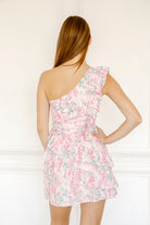 Back view of a woman wearing a pink floral one-shoulder dress against a white background