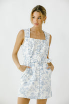 Woman wearing a white sleeveless denim dress with blue toile print and a waist belt against a white background