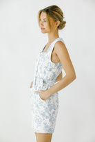 Side view of a woman wearing a white sleeveless denim dress with blue toile print and a waist belt against a white background
