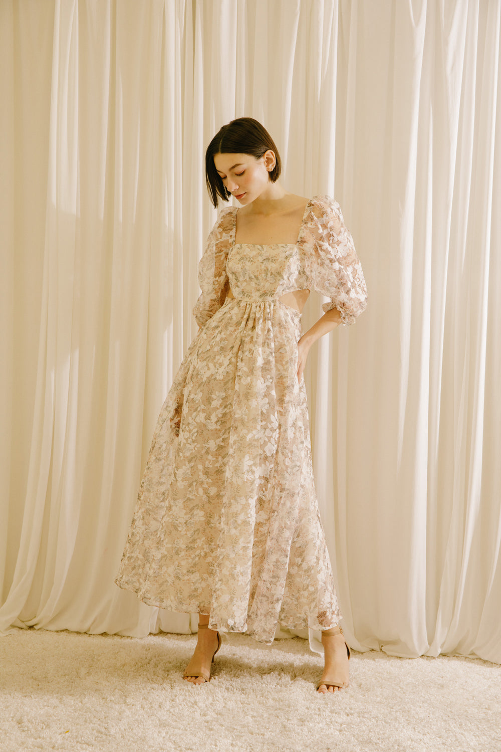 Woman in a floral maxi dress with side cut-outs standing against a beige curtain backdrop