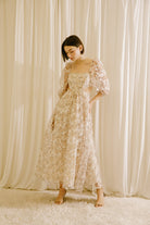 Woman in a floral maxi dress with side cut-outs standing against a beige curtain backdrop