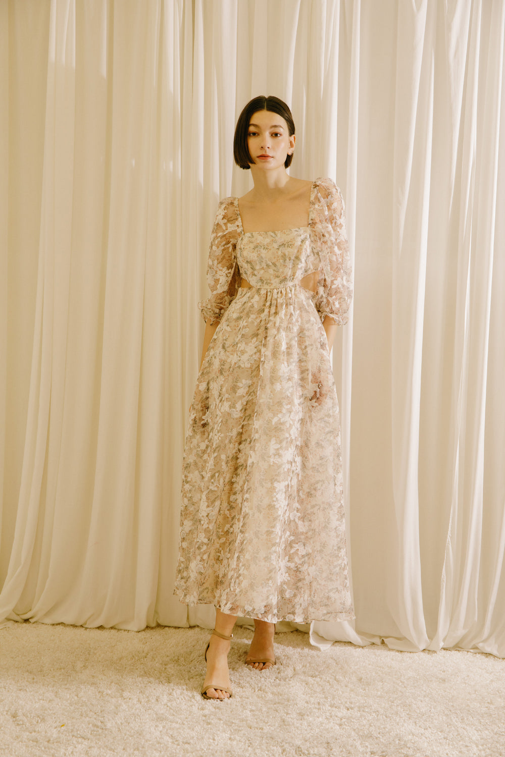 Woman in a floral dress with side cut-outs standing against a beige curtain backdrop