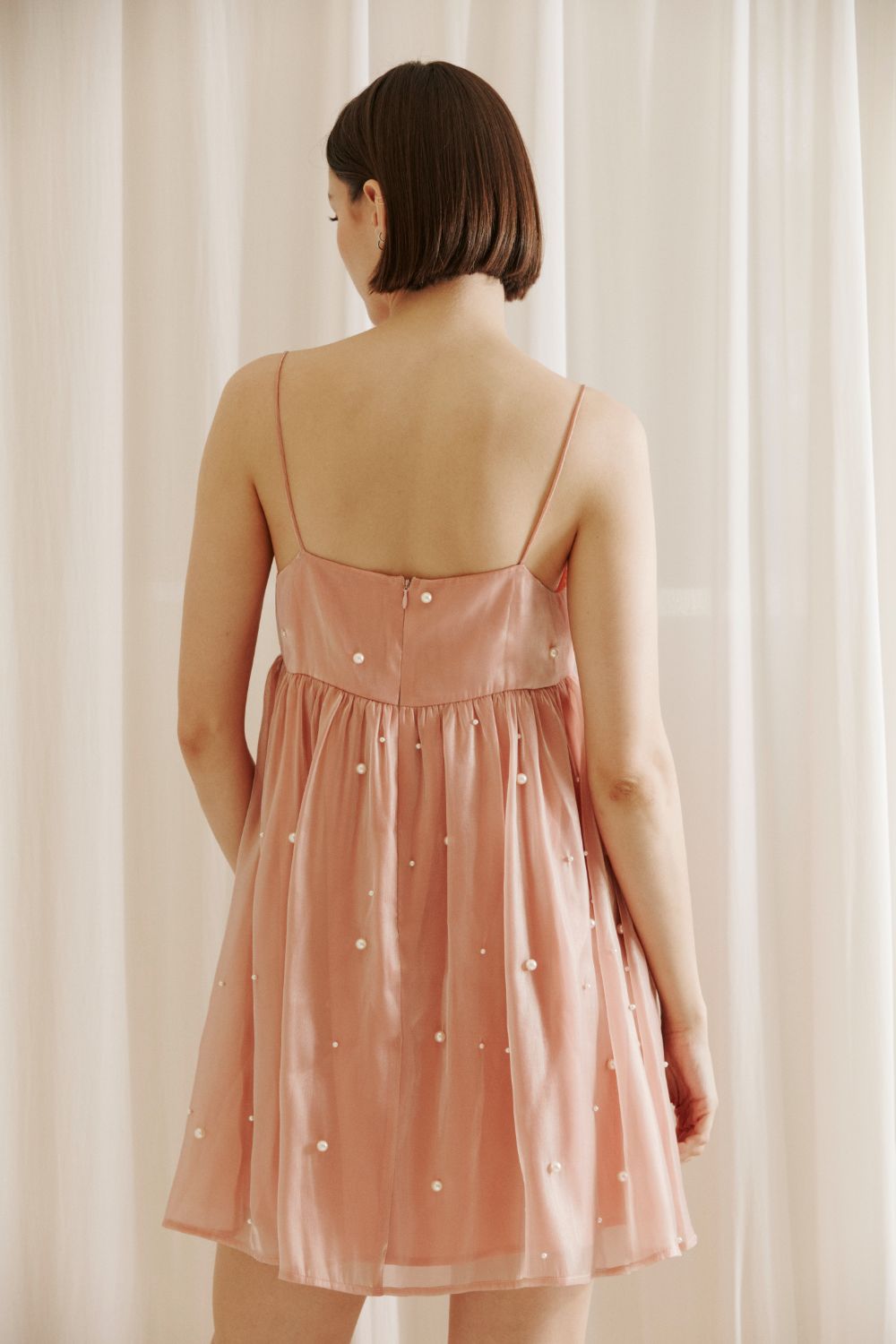 A back view of a person wearing a pink dress with pearl embellishments and an invisible back zipper against a beige curtain background