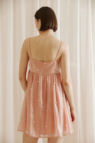 A back view of a person wearing a pink dress with pearl embellishments and an invisible back zipper against a beige curtain background