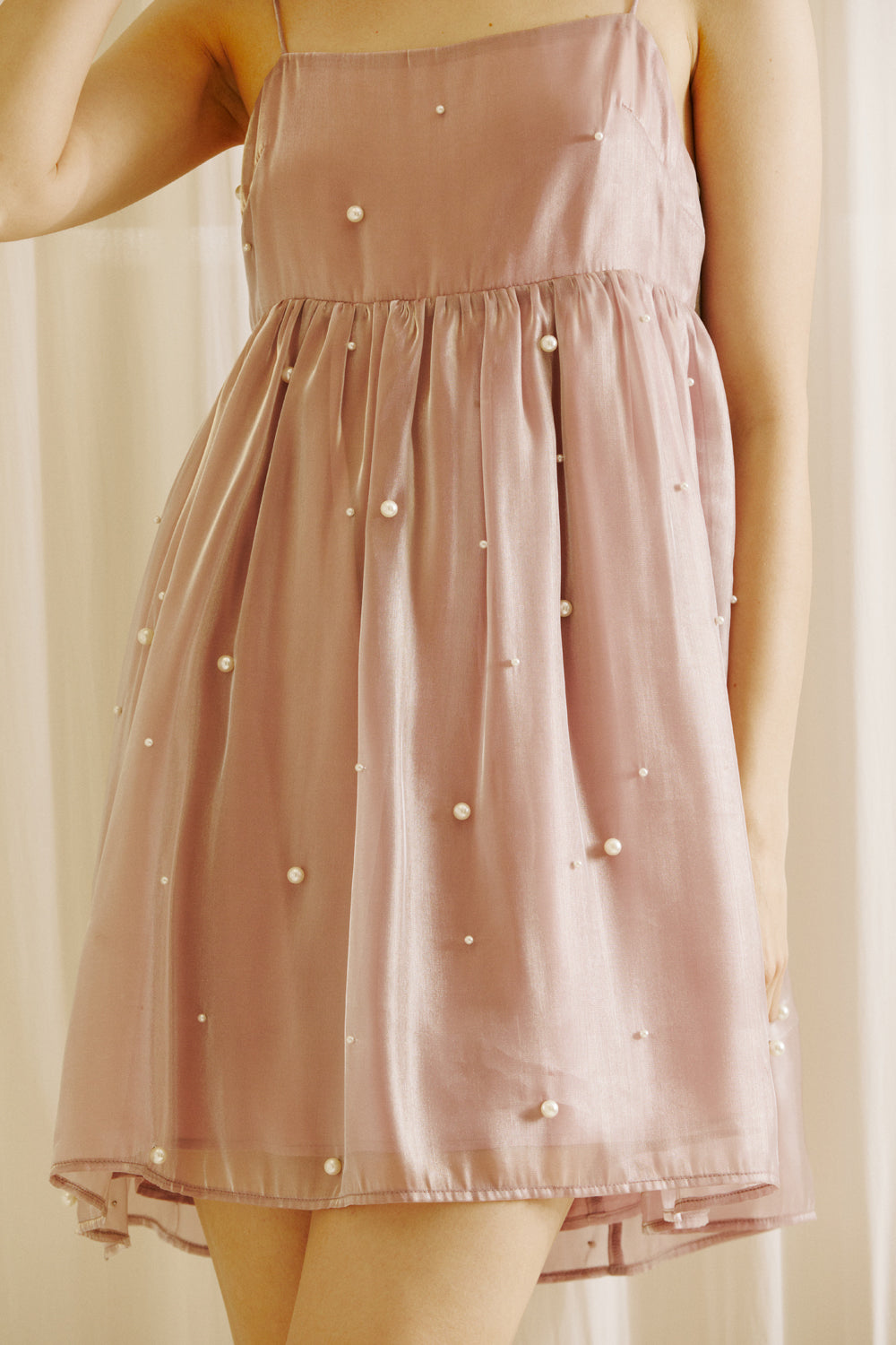 A person wearing a mauve mini dress with pearl embellishments against a beige curtain background