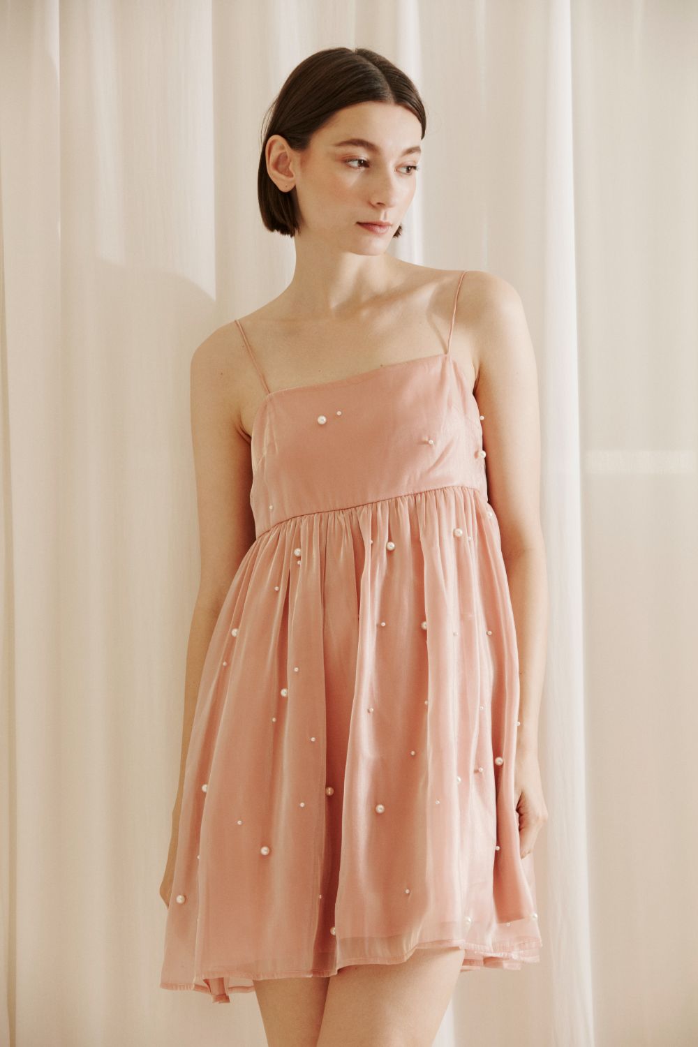 Woman wearing a pink dress with pearl embellishments against a white curtain background
