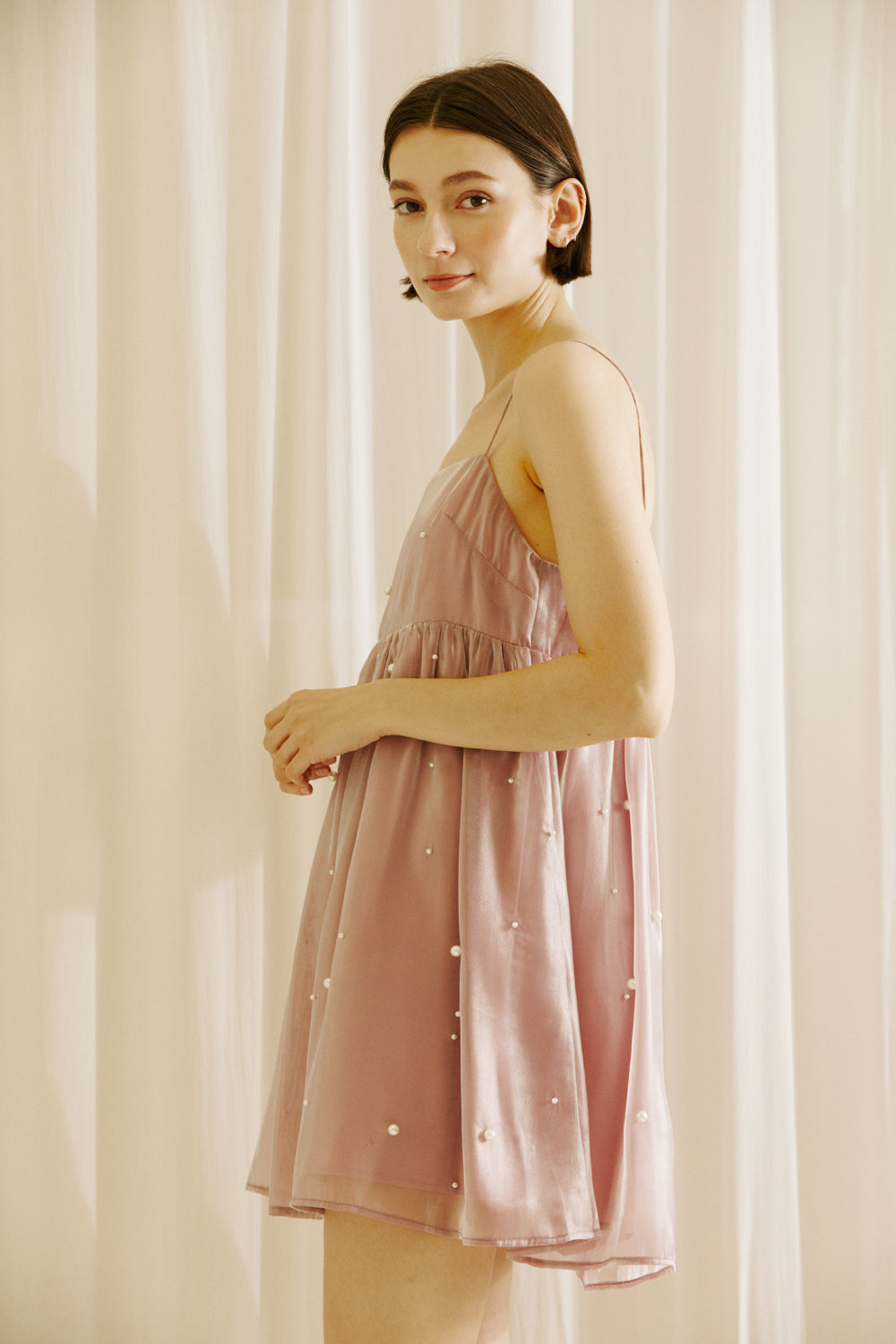 Woman wearing a mauve mini dress with pearl embellishments against a beige curtain background
