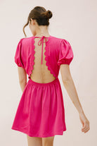 Back view of a woman wearing a bright pink dress with an open back and scalloped hem against a plain background