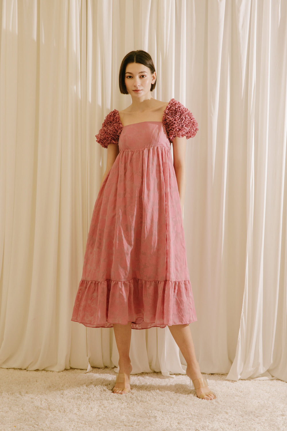 Woman wearing a pink dress with floral sleeves against a beige curtain background