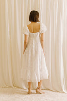 Back view of a woman wearing a white midi dress with statement rosette puff sleeves standing against a beige curtain