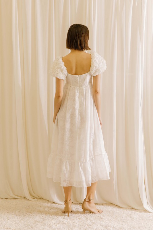 Back view of a woman wearing a white midi dress with statement rosette puff sleeves standing against a beige curtain