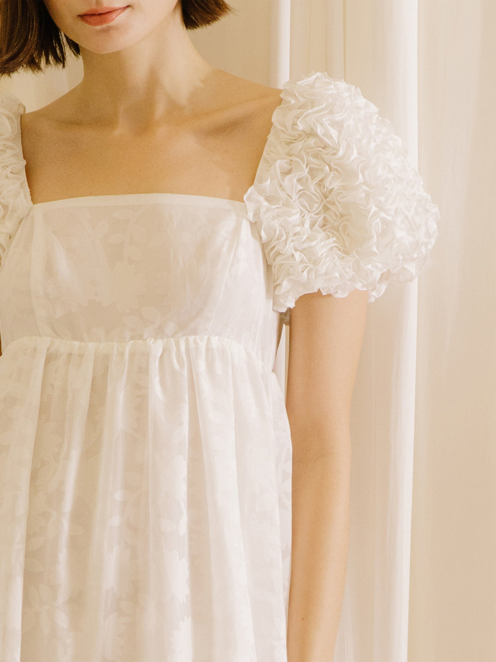 White midi dress with statement rosette puff sleeves standing against a beige curtain