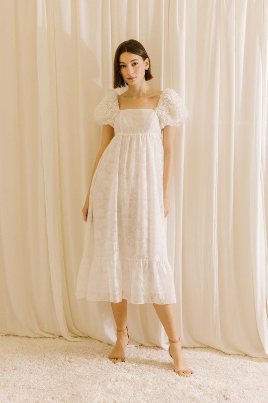 Woman wearing a white midi dress with statement rosette puff sleeves standing against a beige curtain