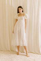 Woman wearing a white midi dress with statement rosette puff sleeves standing against a beige curtain