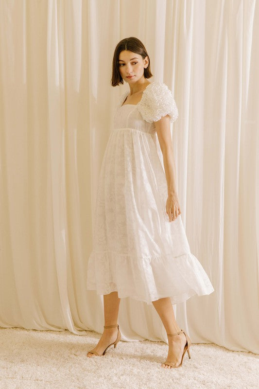 Side view of a woman wearing a white midi dress with statement rosette puff sleeves standing against a beige curtain