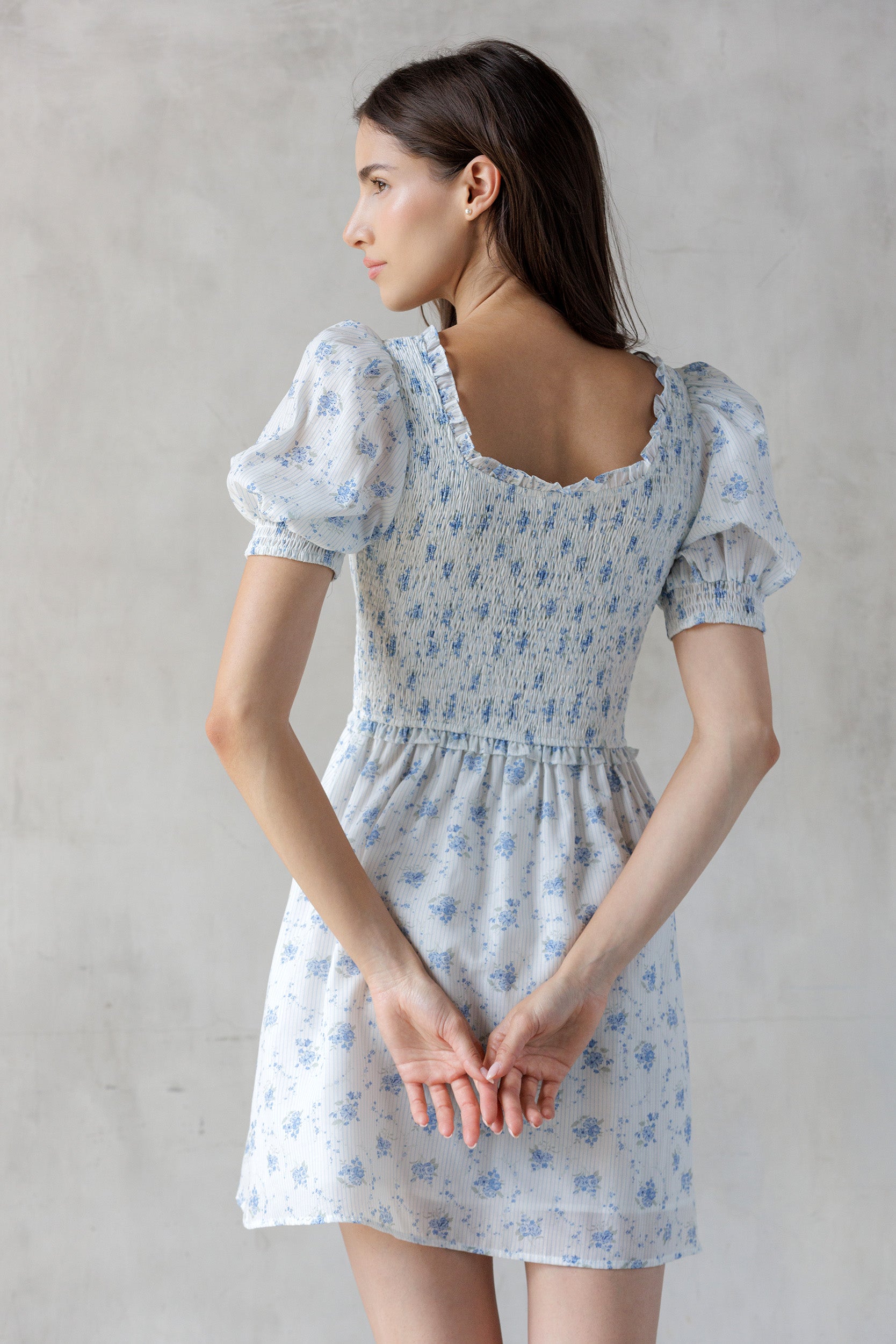 A woman viewed from behind wearing a white and striped dress with a blue floral print  and a smocked bodice against a gray wall.