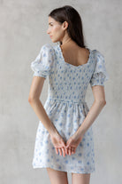 A woman viewed from behind wearing a white and striped dress with a blue floral print  and a smocked bodice against a gray wall.