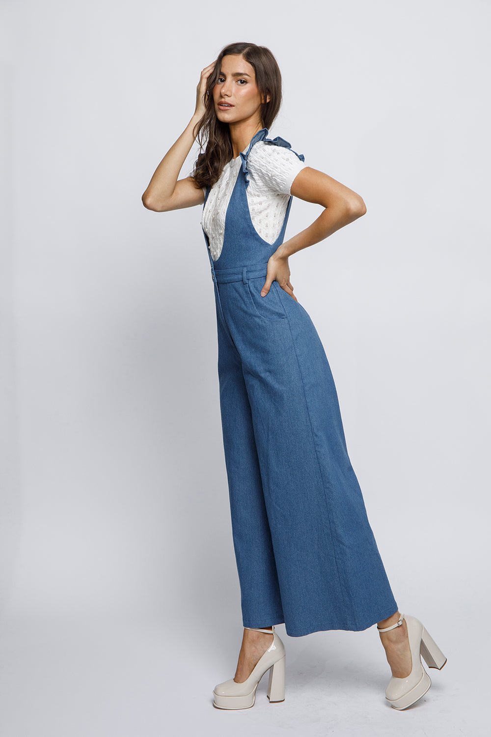 Woman wearing a blue denim jumpsuit with a white top and beige heels on a light gray background