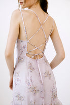 A woman standing in a studio setting, wearing a midi floral dress with ruffles and an open back with a crisscross tie.
