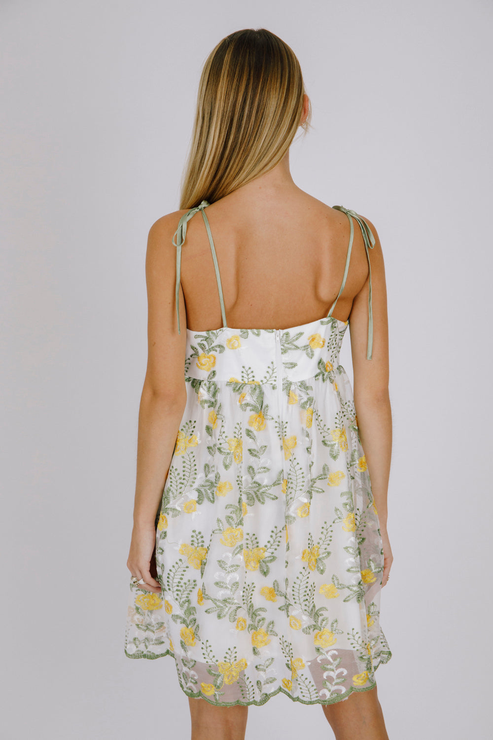 Back view of a woman wearing a floral dress with yellow flowers and self-tie straps on a plain background