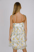 Back view of a woman wearing a floral dress with yellow flowers and self-tie straps on a plain background
