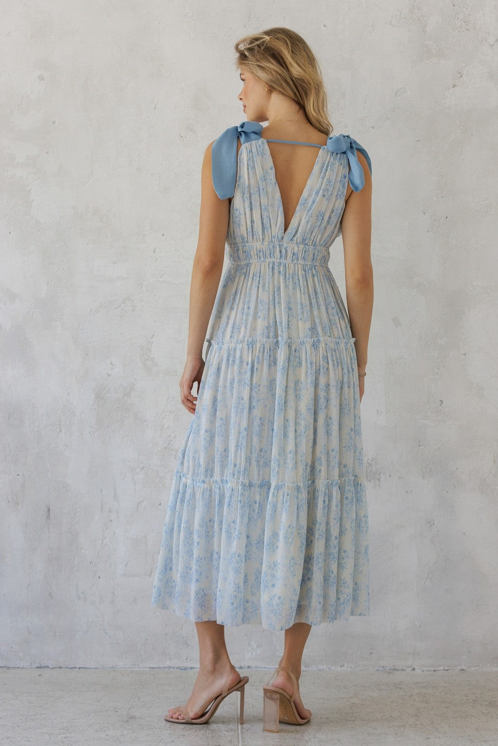 Back of a woman wearing a blue and white striped dress with bow straps against a gray concrete wall.