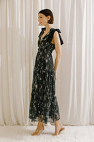 Side view of a model wearing a black and silver metallic floral dress against a white curtain background
