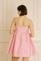 Back view of a woman wearing a pink dress with floral jacquard throughout against a beige wall