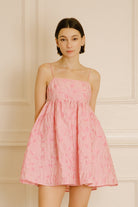 Woman wearing a pink dress with floral jacquard throughout against a beige wall