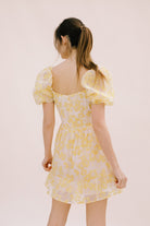Back view of a woman wearing a yellow floral mini dress with an invisible zipper on a light pink background