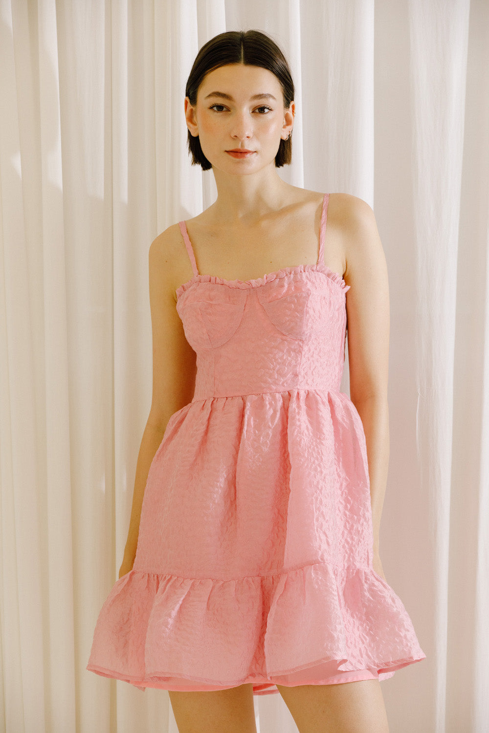 Woman wearing a pink textured dress against a white curtain background
