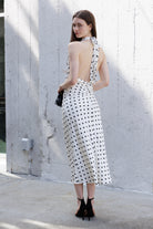 A woman stands outdoors against a textured light gray wall, wearing an ivory halter-neck midi dress with black polka dots and a large open back. She is holding a small black handbag and wearing black ankle-strap heels.