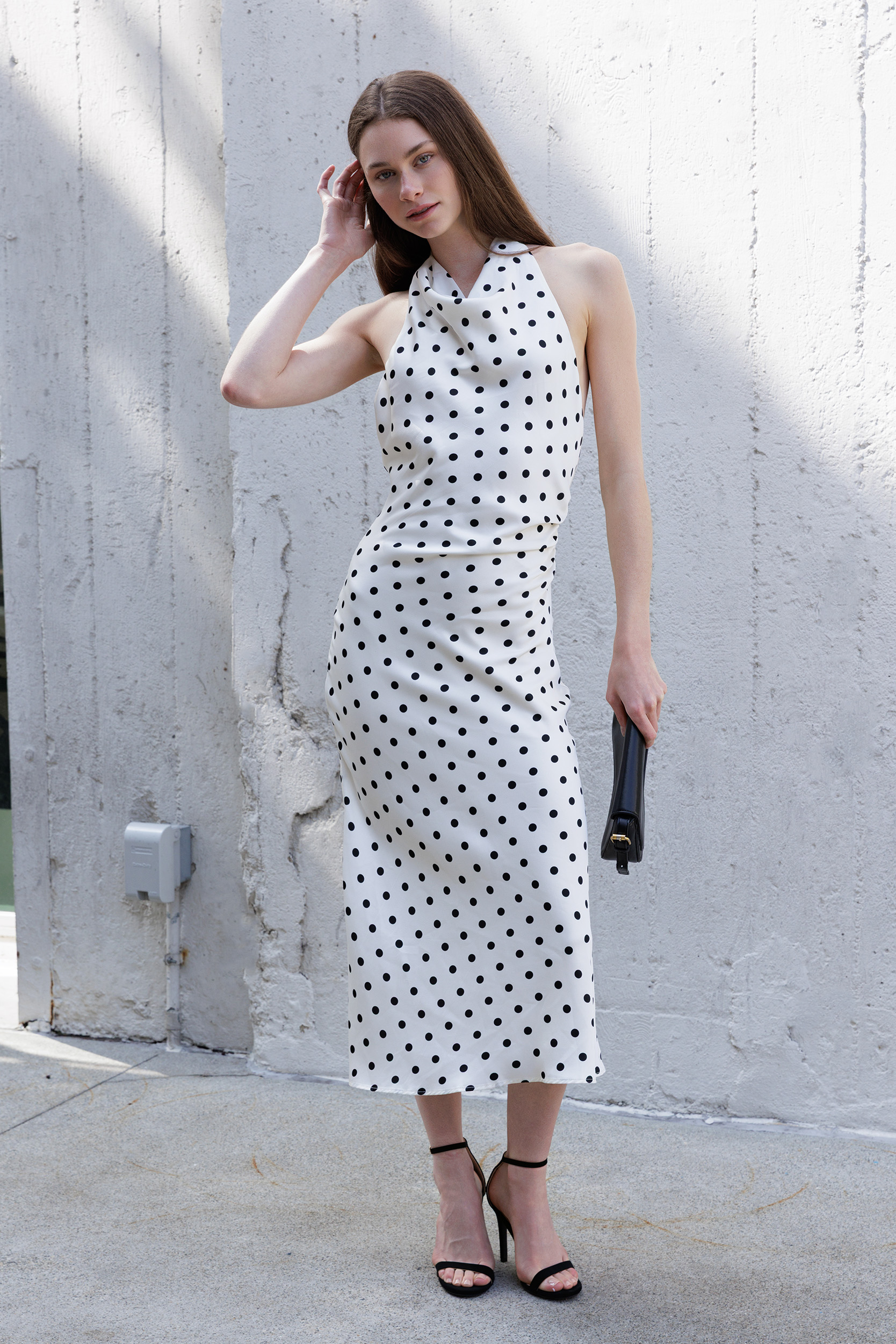 A woman stands against a textured concrete wall wearing a sleeveless ivory midi dress with black polka dots, paired with black heeled sandals and holding a black clutch.
