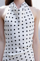 A close-up of a woman wearing a sleeveless halter-neck top or dress in ivory with an allover black polka dot pattern.