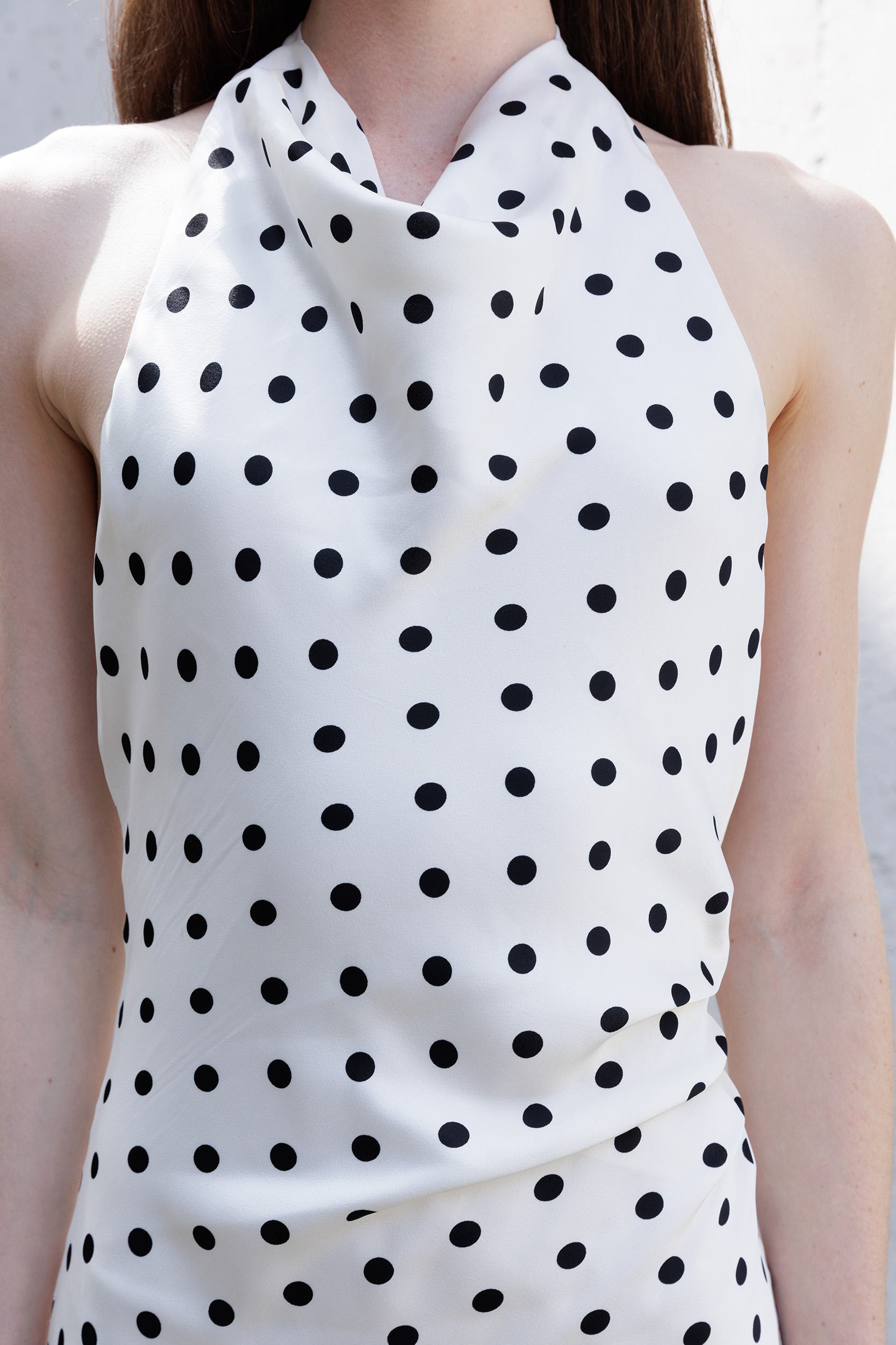 A close-up of a woman wearing a sleeveless halter-neck top or dress in ivory with an allover black polka dot pattern.