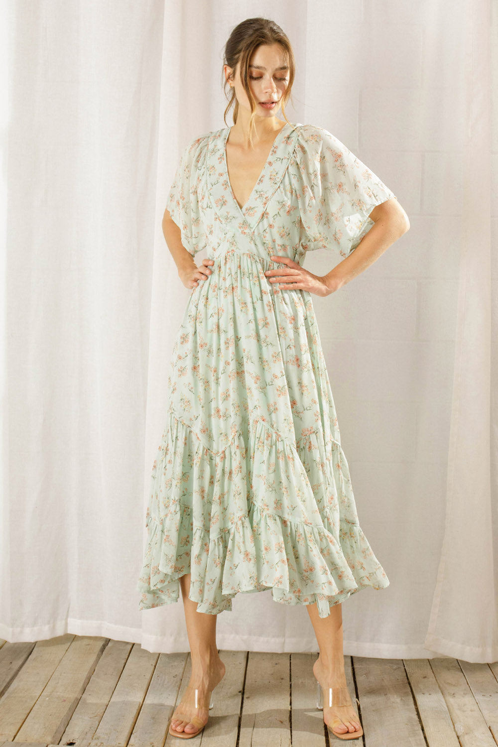 Woman wearing a sage dress with a floral pattern standing against a white curtain background