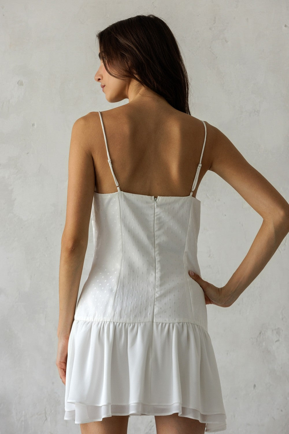 Back of a woman wearing a white dress with thin straps against a plain background