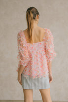 Back view of a woman wearing a pink tulle top with a blue striped skirt against a neutral wall.