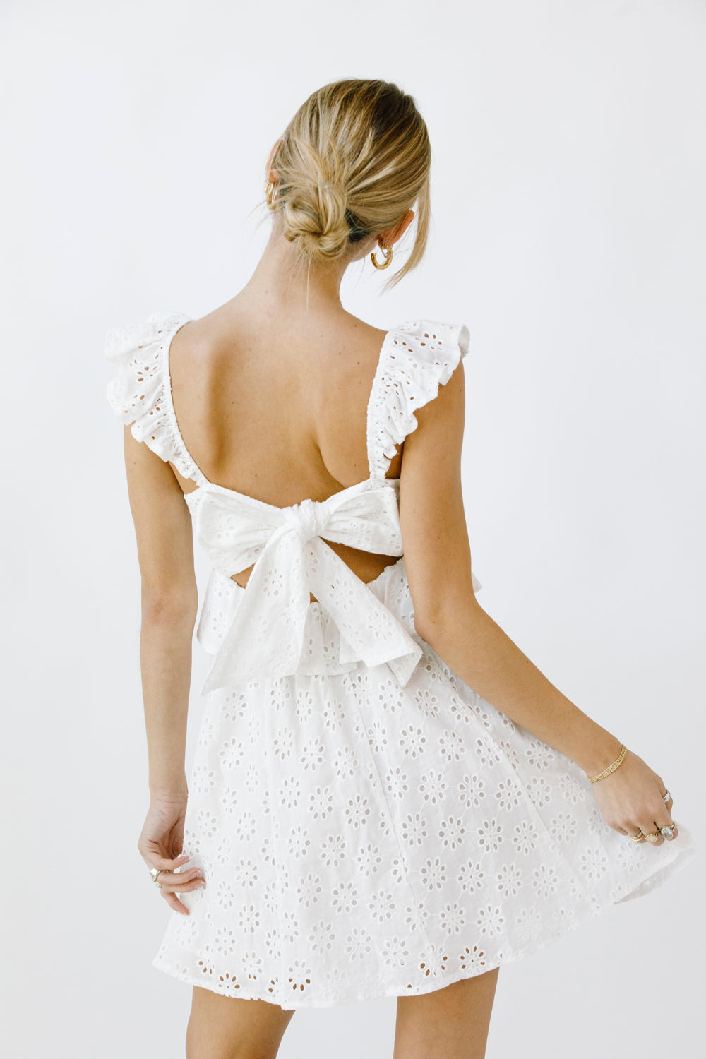 Back view of a woman wearing a white eyelet mini dress with a bow detail on a plain background