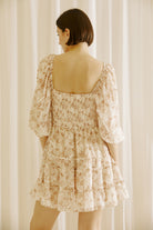 Back view of a model wearing a beige and brown 0dress with a classy daisy print and standing against a white curtain