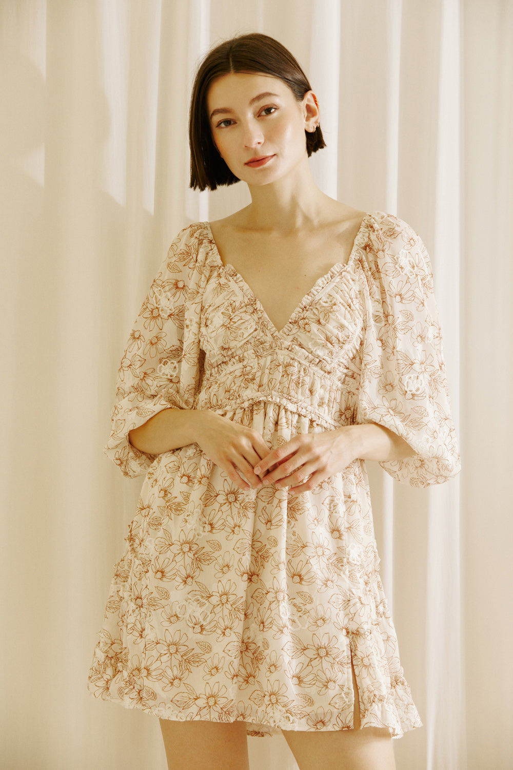 Woman wearing a beige and brown 0dress with a classy daisy print and standing against a white curtain