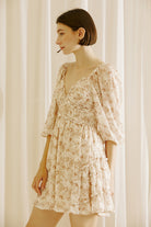 Side view of a woman wearing a beige and brown 0dress with a classy daisy print and standing against a white curtain