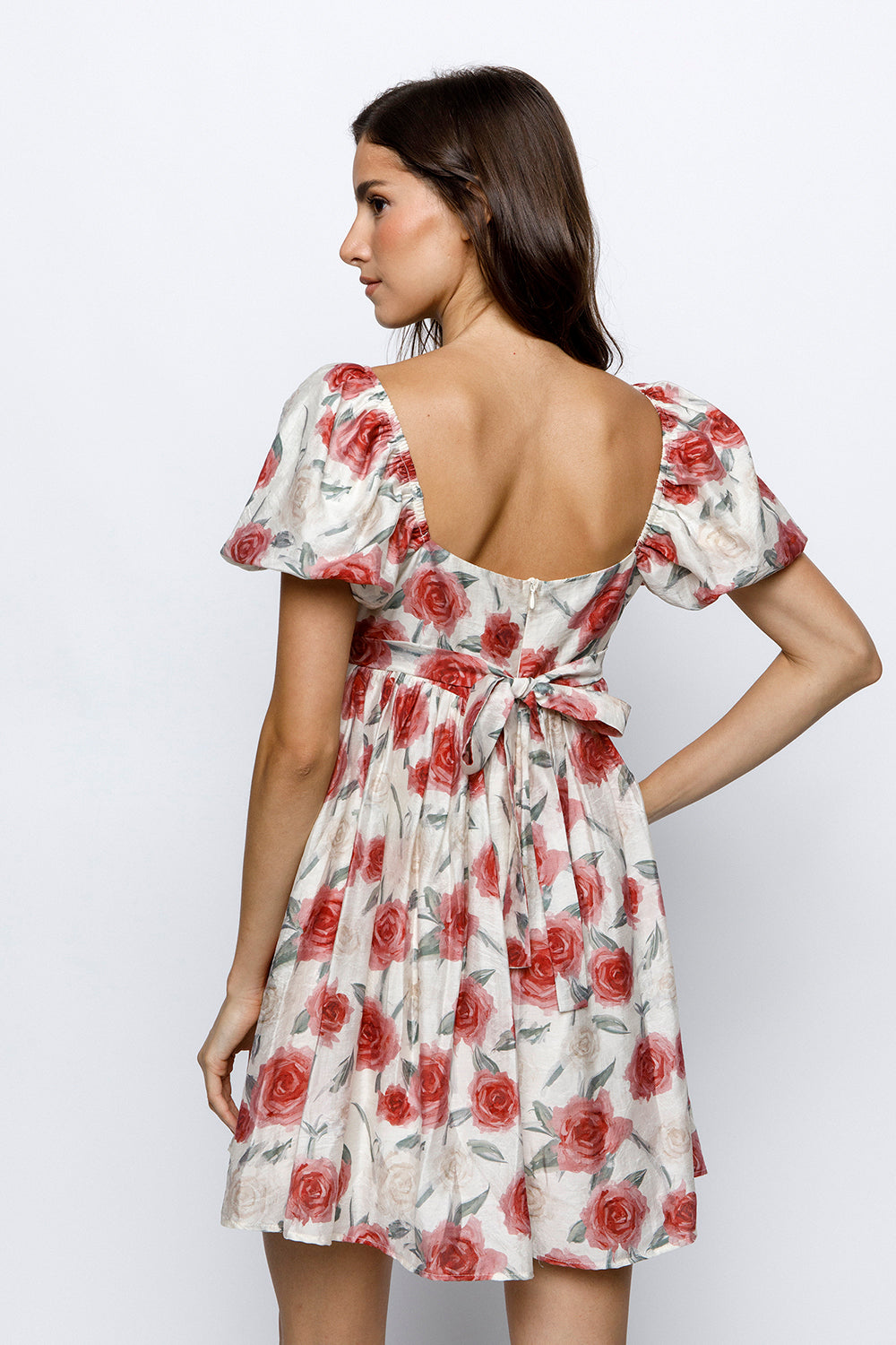 Back view of a woman wearing an ivory  dress with red rose pattern and a self-tie bow a white background